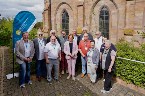 12 Digital-Botschafterinnen und -Botschafter stehen in einem Halbkreis um Ministerpräsidentin Malu Dreyer. Alle lachen in die Kamera. Die Sonne scheint. Im Hintergrund ist die Kirche in Harxheim Zellertal zu sehen.